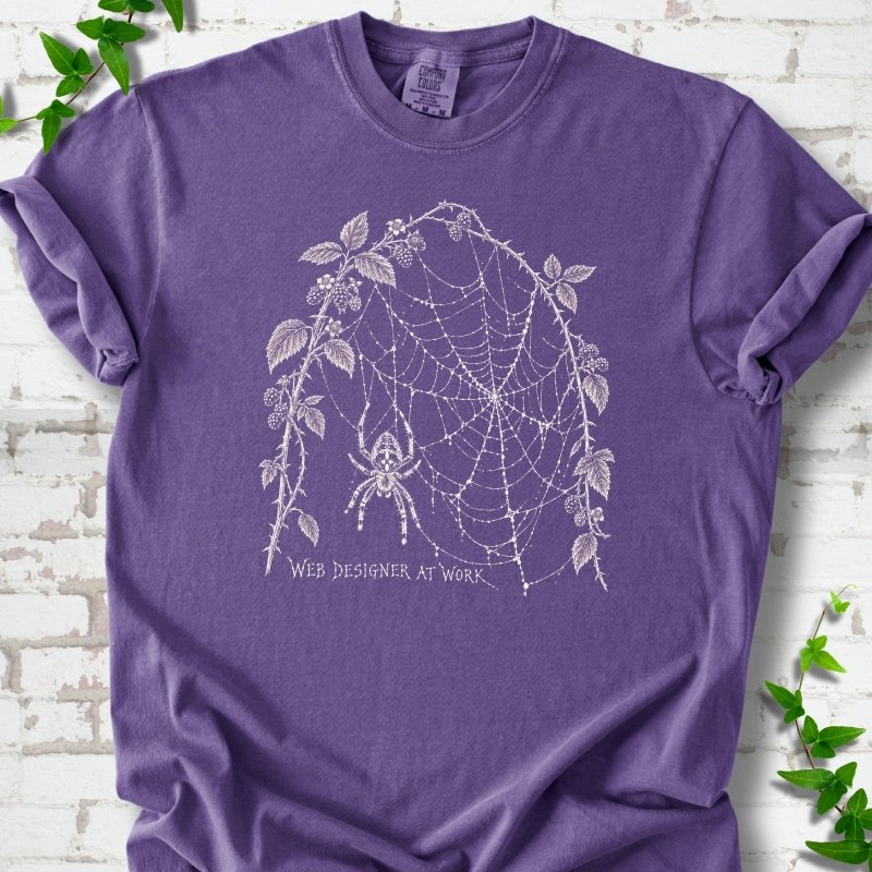 Grape Comfort Colors® T-shirt laid flat, showing a cream spider and intricate web framed by trailing leaves, with “Web designer at work” below.