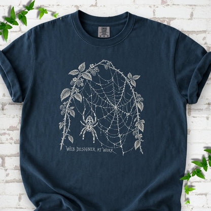Midnight Comfort Colors® T-shirt laid flat, showing a cream spider and intricate web framed by trailing leaves, with “Web designer at work” below.