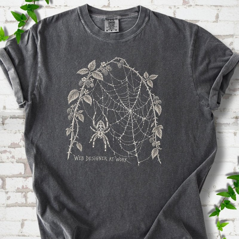 Pepper Comfort Colors® T-shirt laid flat, showing a cream spider and intricate web framed by trailing leaves, with “Web designer at work” below.