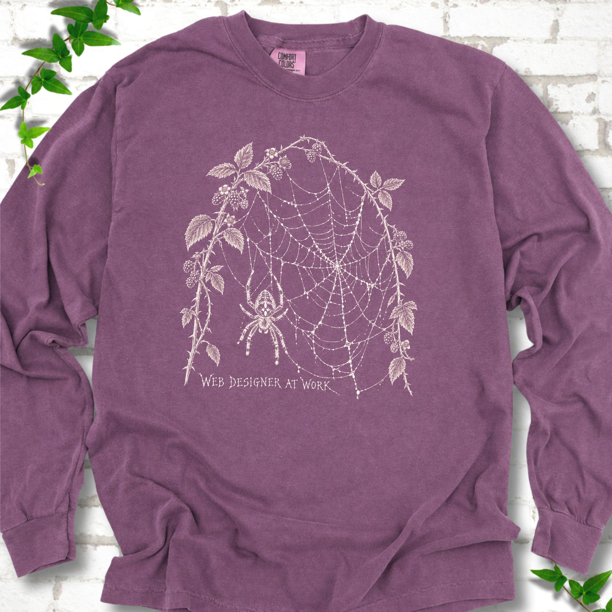 Berry Comfort Colors® long sleeve T-shirt laid flat, showing a cream spider and intricate web framed by trailing leaves, with “Web designer at work” below.