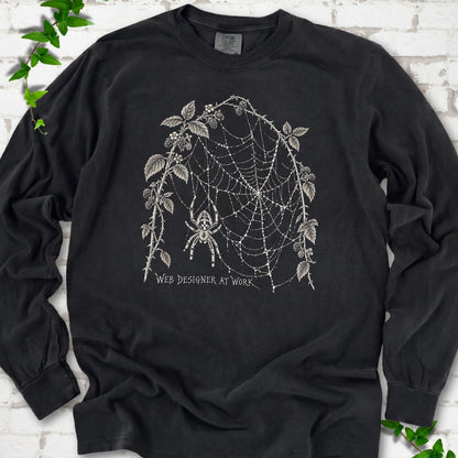 Black Comfort Colors® long sleeve T-shirt laid flat, showing a cream spider and intricate web framed by trailing leaves, with “Web designer at work” below.