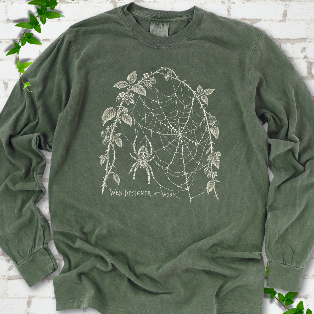 Hemp Comfort Colors® long sleeve T-shirt laid flat, showing a cream spider and intricate web framed by trailing leaves, with “Web designer at work” below.