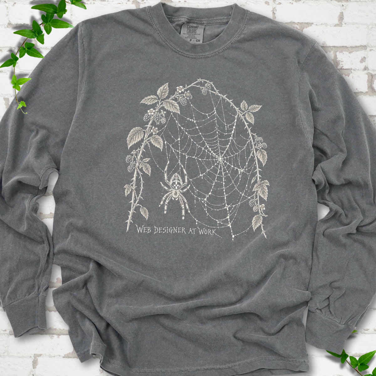 Pepper Comfort Colors® long sleeve T-shirt laid flat, showing a cream spider and intricate web framed by trailing leaves, with “Web designer at work” below.