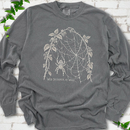 Pepper Comfort Colors® long sleeve T-shirt laid flat, showing a cream spider and intricate web framed by trailing leaves, with “Web designer at work” below.