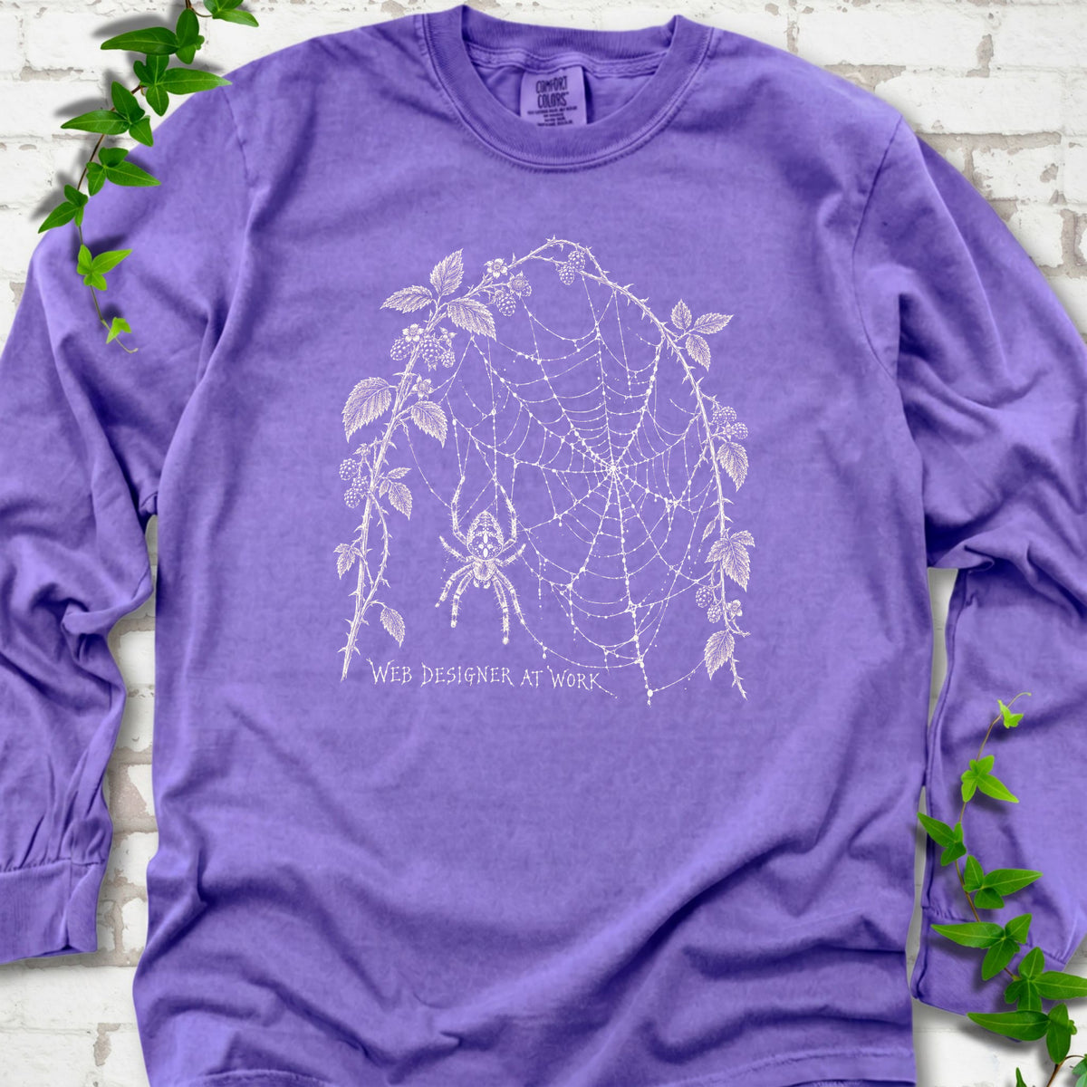 Violet Comfort Colors® long sleeve T-shirt laid flat, showing a cream spider and intricate web framed by trailing leaves, with “Web designer at work” below.