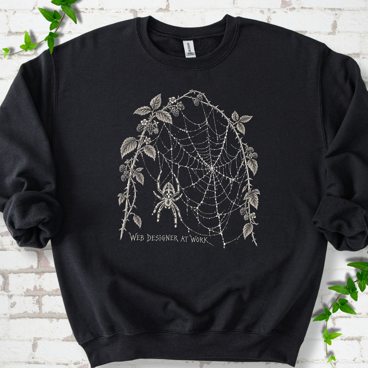 Black classic sweatshirt laid flat, showing a cream spider and intricate web framed by trailing leaves, with “Web designer at work” below.