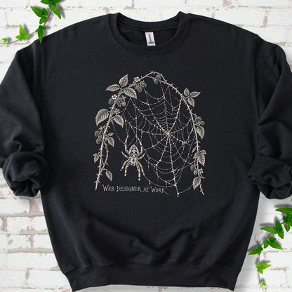Black classic sweatshirt laid flat, showing a cream spider and intricate web framed by trailing leaves, with “Web designer at work” below.