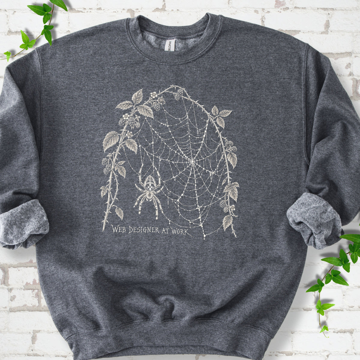 Dark Heather classic sweatshirt laid flat, showing a cream spider and intricate web framed by trailing leaves, with “Web designer at work” below.