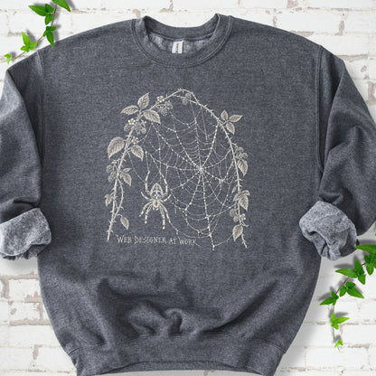 Dark Heather classic sweatshirt laid flat, showing a cream spider and intricate web framed by trailing leaves, with “Web designer at work” below.