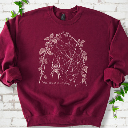 Maroon classic sweatshirt laid flat, showing a cream spider and intricate web framed by trailing leaves, with “Web designer at work” below.