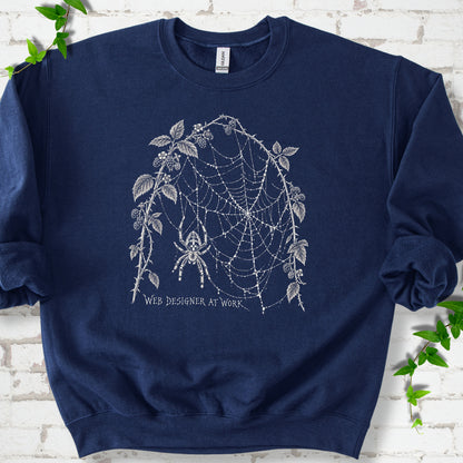 Navy classic sweatshirt laid flat, showing a cream spider and intricate web framed by trailing leaves, with “Web designer at work” below.