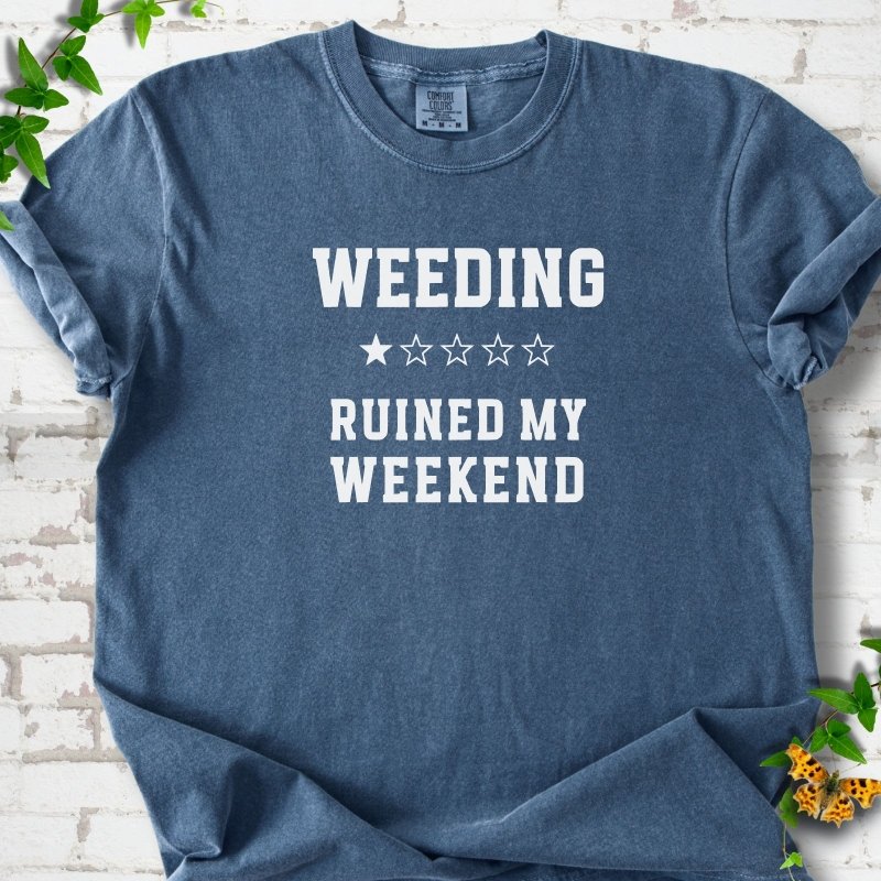 Denim short sleeve T-shirt laid flat on white brick with greenery, showing cream 'Weeding Ruined My Weekend' lettering and a one-star rating on the chest.