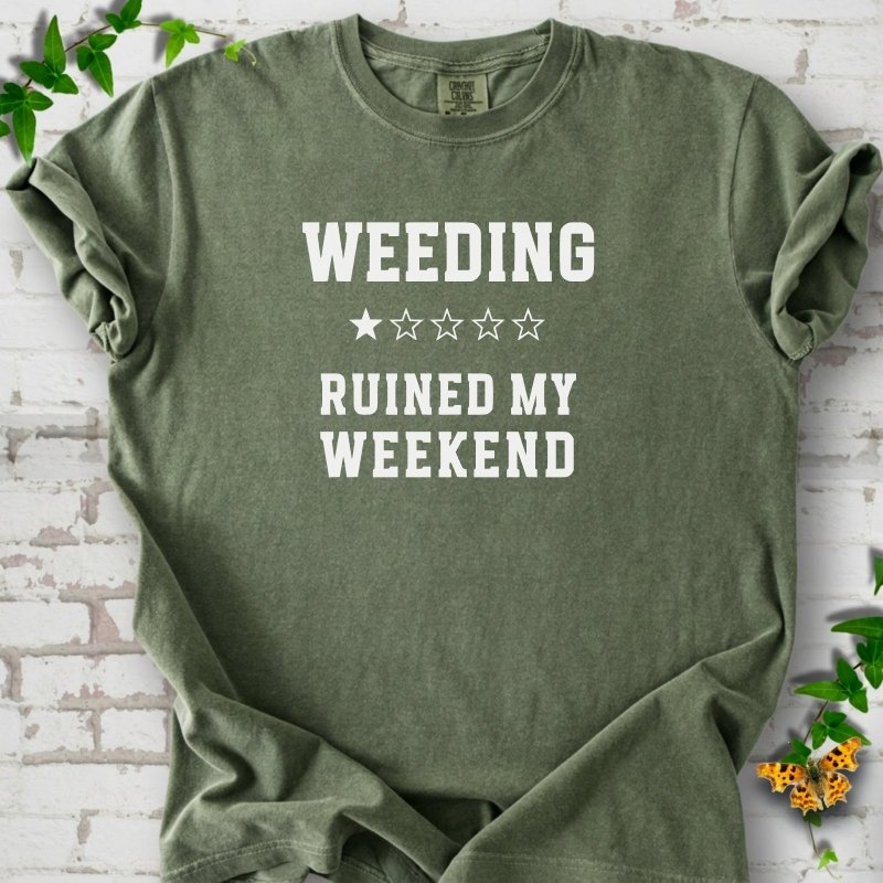 Moss short sleeve T-shirt laid flat on white brick with greenery, showing cream 'Weeding Ruined My Weekend' lettering and a one-star rating on the chest.
