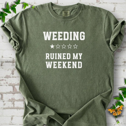 Moss short sleeve T-shirt laid flat on white brick with greenery, showing cream 'Weeding Ruined My Weekend' lettering and a one-star rating on the chest.