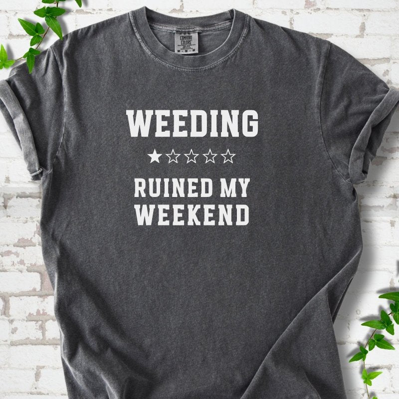 Pepper short sleeve T-shirt laid flat on white brick with greenery, showing cream 'Weeding Ruined My Weekend' lettering and a one-star rating on the chest.