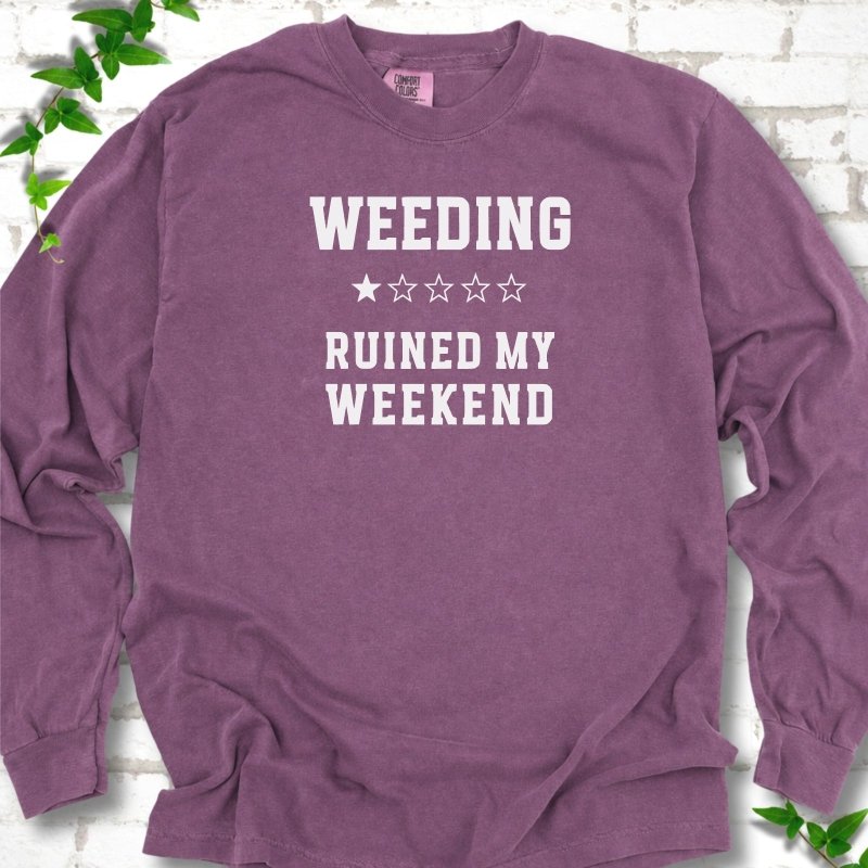 Berry long sleeve T-shirt laid flat on white brick with greenery, showing cream 'Weeding Ruined My Weekend' lettering and a one-star rating on the chest.