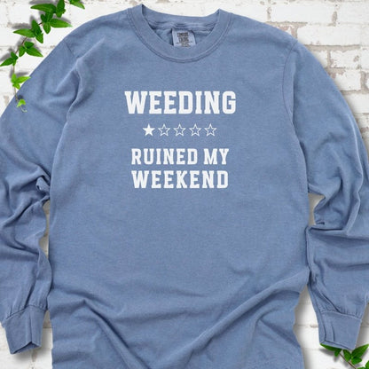Blue Jean long sleeve T-shirt laid flat on white brick with greenery, showing cream 'Weeding Ruined My Weekend' lettering and a one-star rating on the chest.
