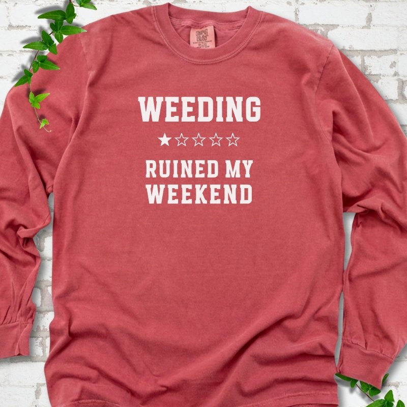 Crimson long sleeve T-shirt laid flat on white brick with greenery, showing cream 'Weeding Ruined My Weekend' lettering and a one-star rating on the chest.