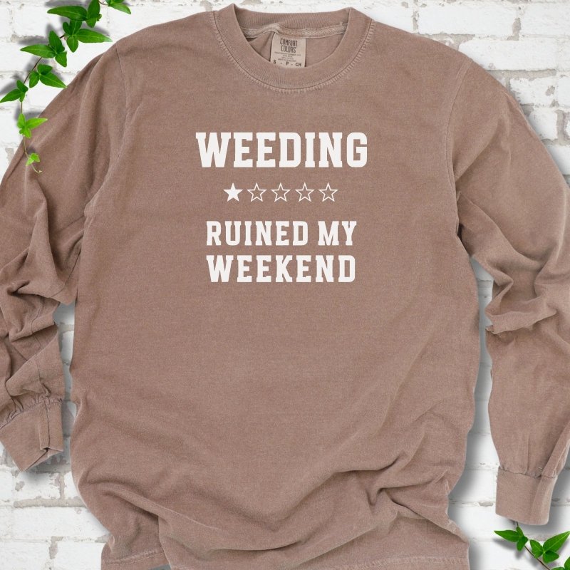 Espresso long sleeve T-shirt laid flat on white brick with greenery, showing cream 'Weeding Ruined My Weekend' lettering and a one-star rating on the chest.
