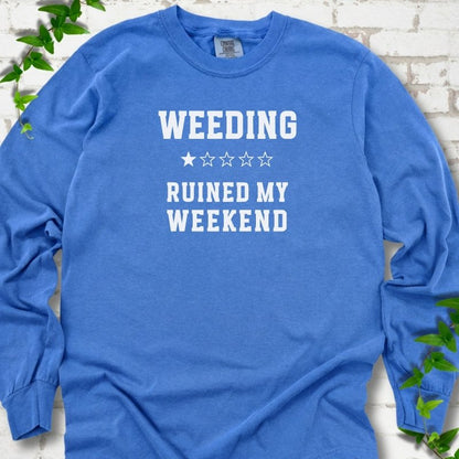 Flo Blue long sleeve T-shirt laid flat on white brick with greenery, showing cream 'Weeding Ruined My Weekend' lettering and a one-star rating on the chest.