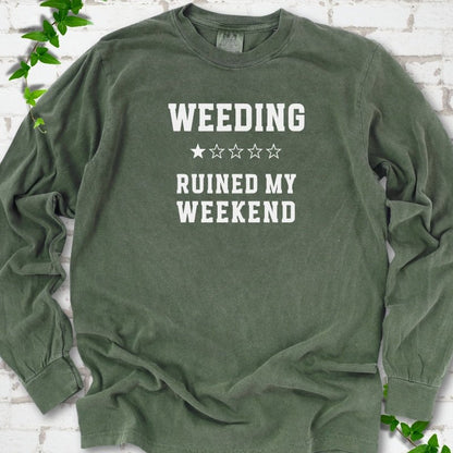 Hemp long sleeve T-shirt laid flat on white brick with greenery, showing cream 'Weeding Ruined My Weekend' lettering and a one-star rating on the chest.
