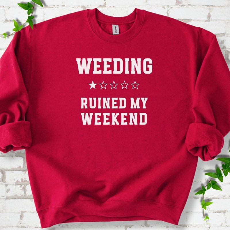 Cardinal Red crewneck sweatshirt laid flat on white brick with greenery, showing cream 'Weeding Ruined My Weekend' lettering and a one-star rating on the chest.