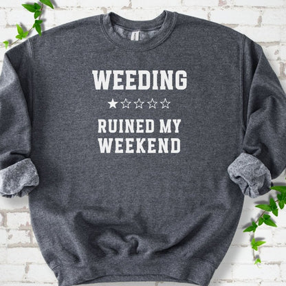 Dark Heather crewneck sweatshirt laid flat on white brick with greenery, showing cream 'Weeding Ruined My Weekend' lettering and a one-star rating on the chest.
