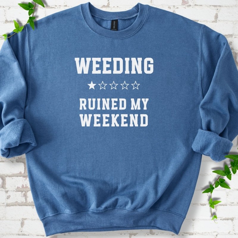 Indigo Blue crewneck sweatshirt laid flat on white brick with greenery, showing cream 'Weeding Ruined My Weekend' lettering and a one-star rating on the chest.