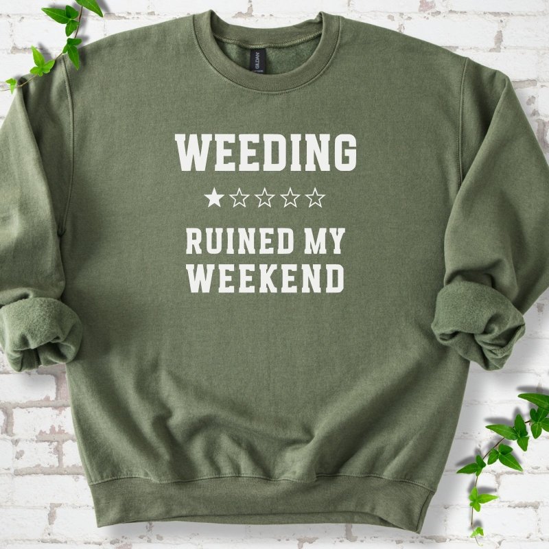 Military Green crewneck sweatshirt laid flat on white brick with greenery, showing cream 'Weeding Ruined My Weekend' lettering and a one-star rating on the chest.