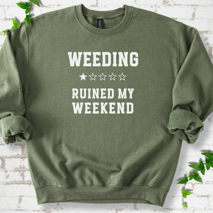 Military Green crewneck sweatshirt laid flat on white brick with greenery, showing cream 'Weeding Ruined My Weekend' lettering and a one-star rating on the chest.
