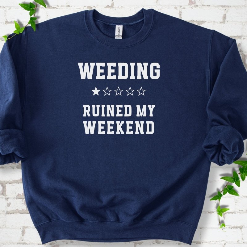Navy crewneck sweatshirt laid flat on white brick with greenery, showing cream 'Weeding Ruined My Weekend' lettering and a one-star rating on the chest.