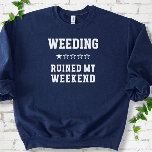 Navy crewneck sweatshirt laid flat on white brick with greenery, showing cream 'Weeding Ruined My Weekend' lettering and a one-star rating on the chest.