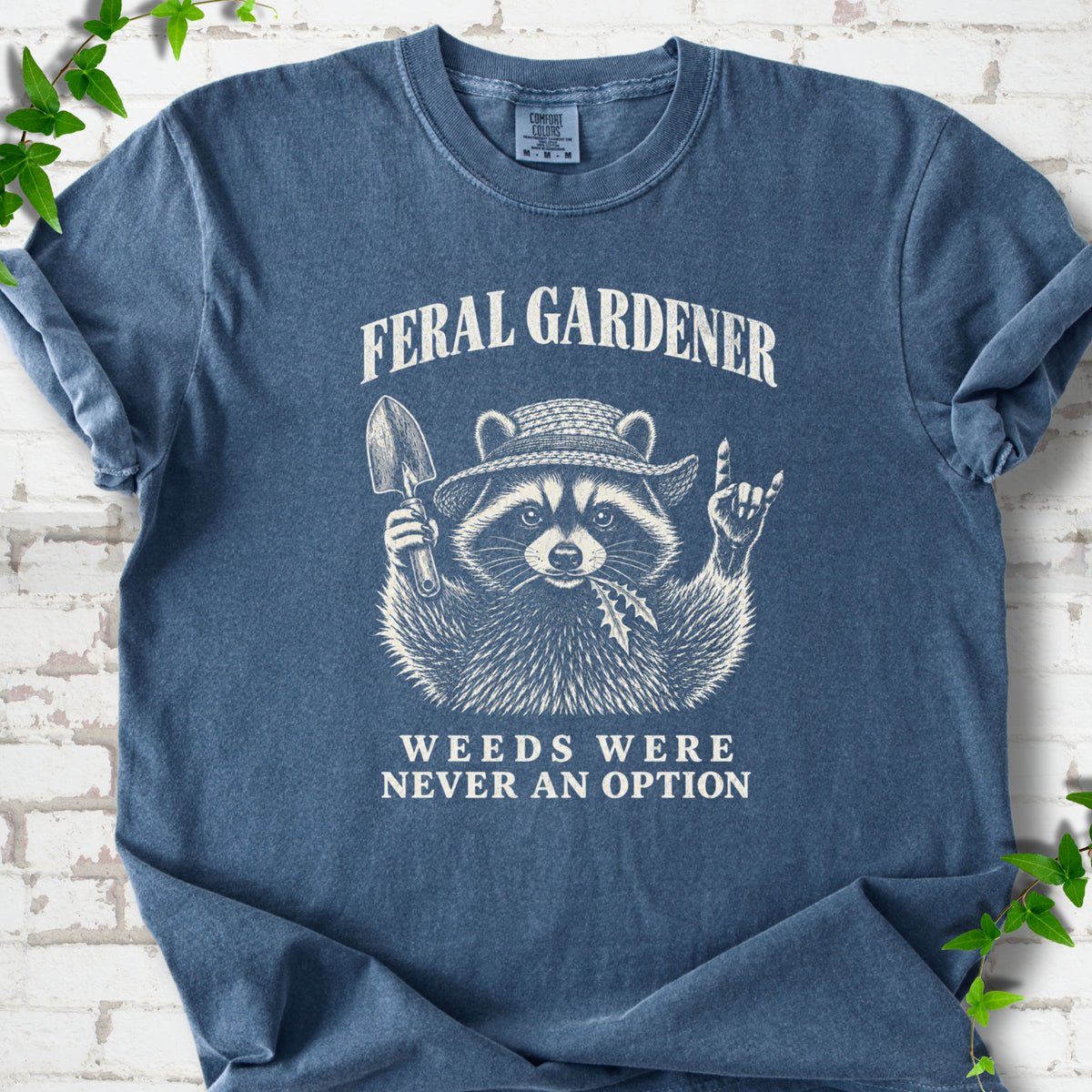 Denim T-shirt laid flat, showing a cream raccoon in a straw hat holding a trowel, with "Feral Gardener" and "Weeds Were Never an Option" lettering.
