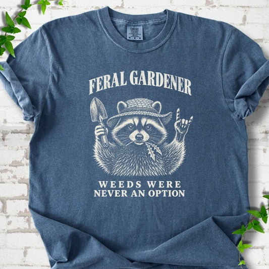 Denim T-shirt laid flat, showing a cream raccoon in a straw hat holding a trowel, with "Feral Gardener" and "Weeds Were Never an Option" lettering.