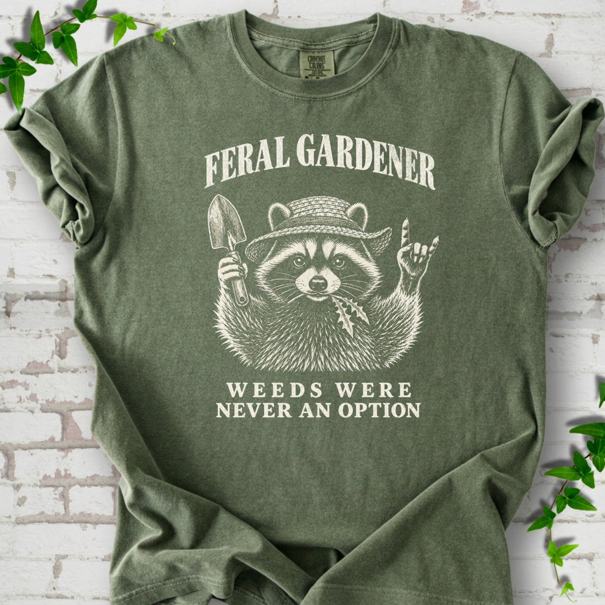 Moss T-shirt laid flat, showing a cream raccoon in a straw hat holding a trowel, with "Feral Gardener" and "Weeds Were Never an Option" lettering.