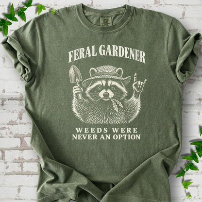 Moss T-shirt laid flat, showing a cream raccoon in a straw hat holding a trowel, with "Feral Gardener" and "Weeds Were Never an Option" lettering.