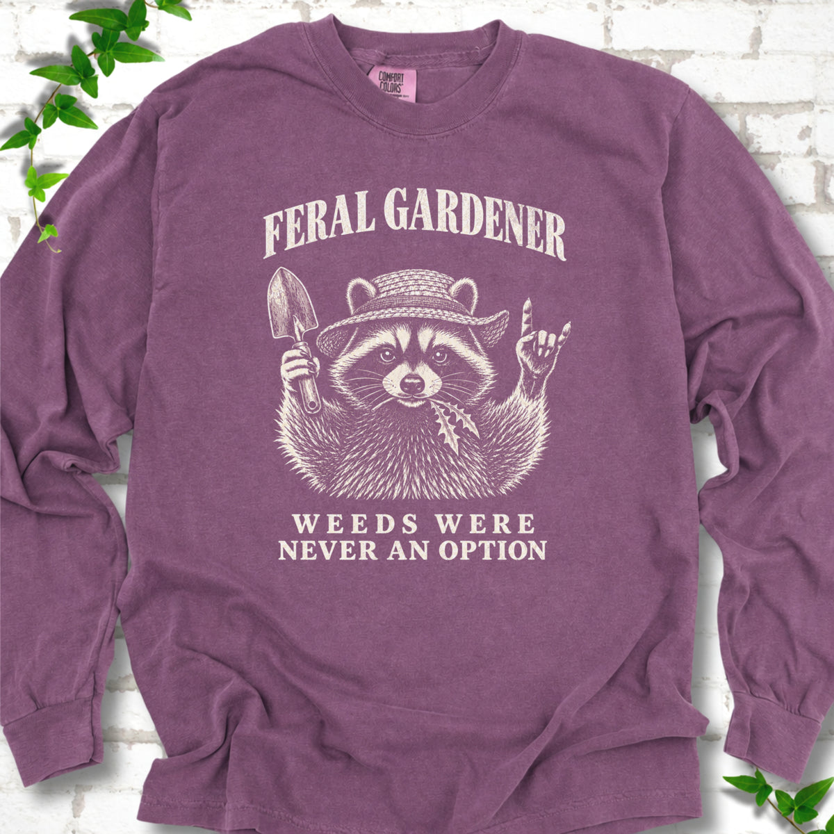 Berry long sleeve tee laid flat, showing a cream raccoon in a straw hat holding a trowel, with "Feral Gardener" and "Weeds Were Never an Option" lettering.