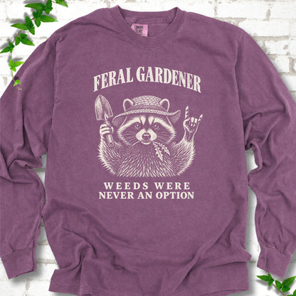 Berry long sleeve tee laid flat, showing a cream raccoon in a straw hat holding a trowel, with "Feral Gardener" and "Weeds Were Never an Option" lettering.
