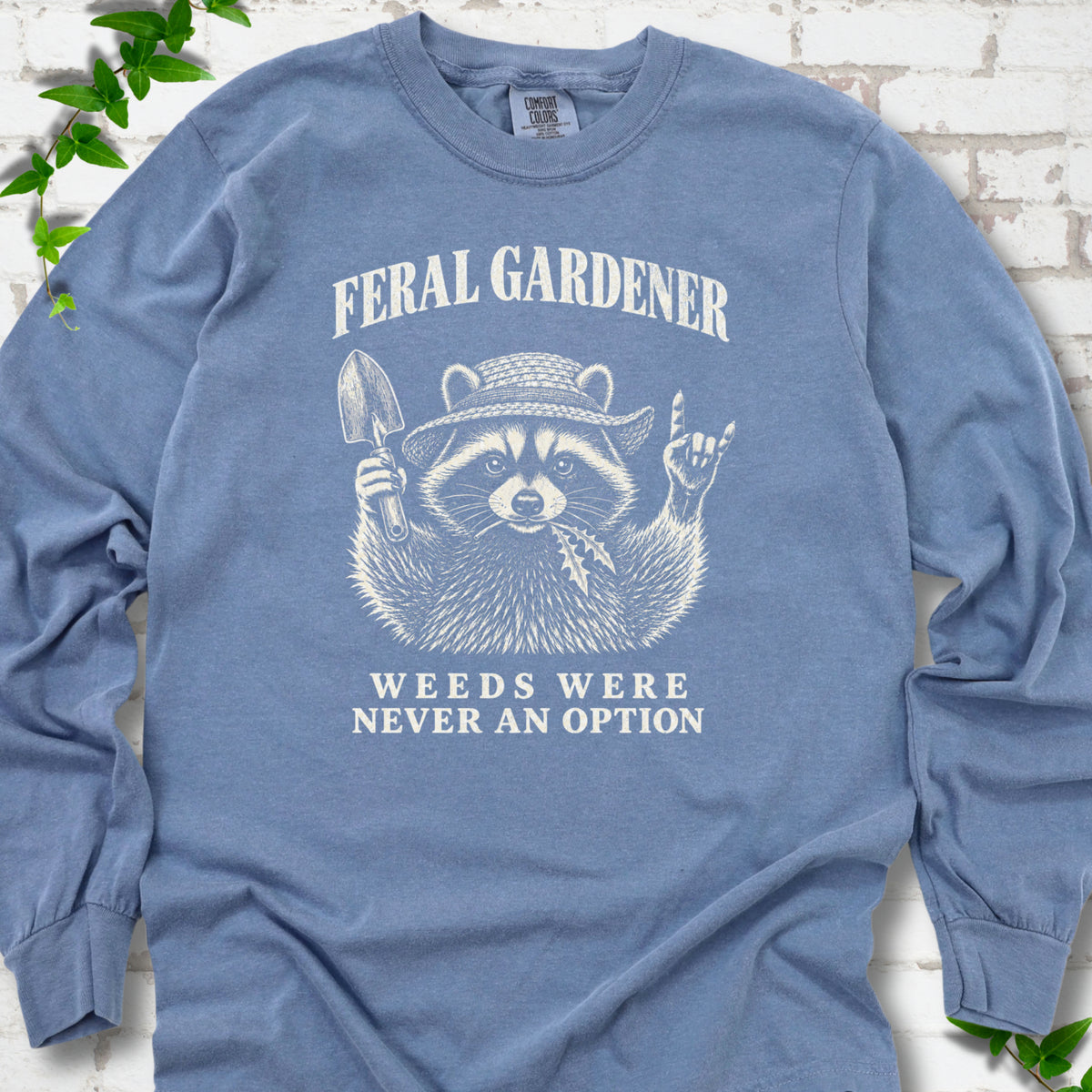 Blue Jean long sleeve tee laid flat, showing a cream raccoon in a straw hat holding a trowel, with "Feral Gardener" and "Weeds Were Never an Option" lettering.