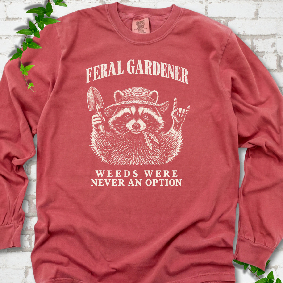 Crimson long sleeve tee laid flat, showing a cream raccoon in a straw hat holding a trowel, with "Feral Gardener" and "Weeds Were Never an Option" lettering.