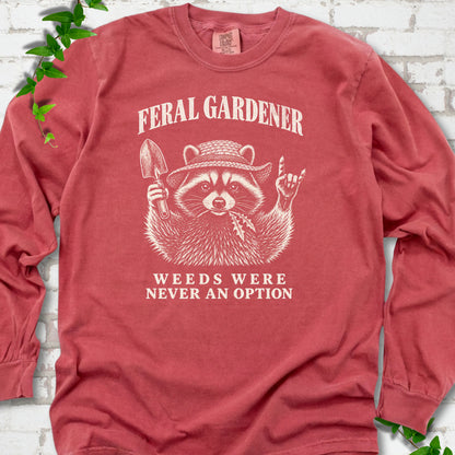 Crimson long sleeve tee laid flat, showing a cream raccoon in a straw hat holding a trowel, with "Feral Gardener" and "Weeds Were Never an Option" lettering.