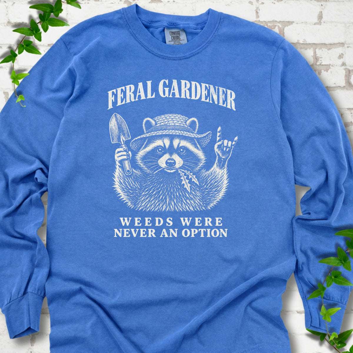 Flo Blue long sleeve tee laid flat, showing a cream raccoon in a straw hat holding a trowel, with "Feral Gardener" and "Weeds Were Never an Option" lettering.