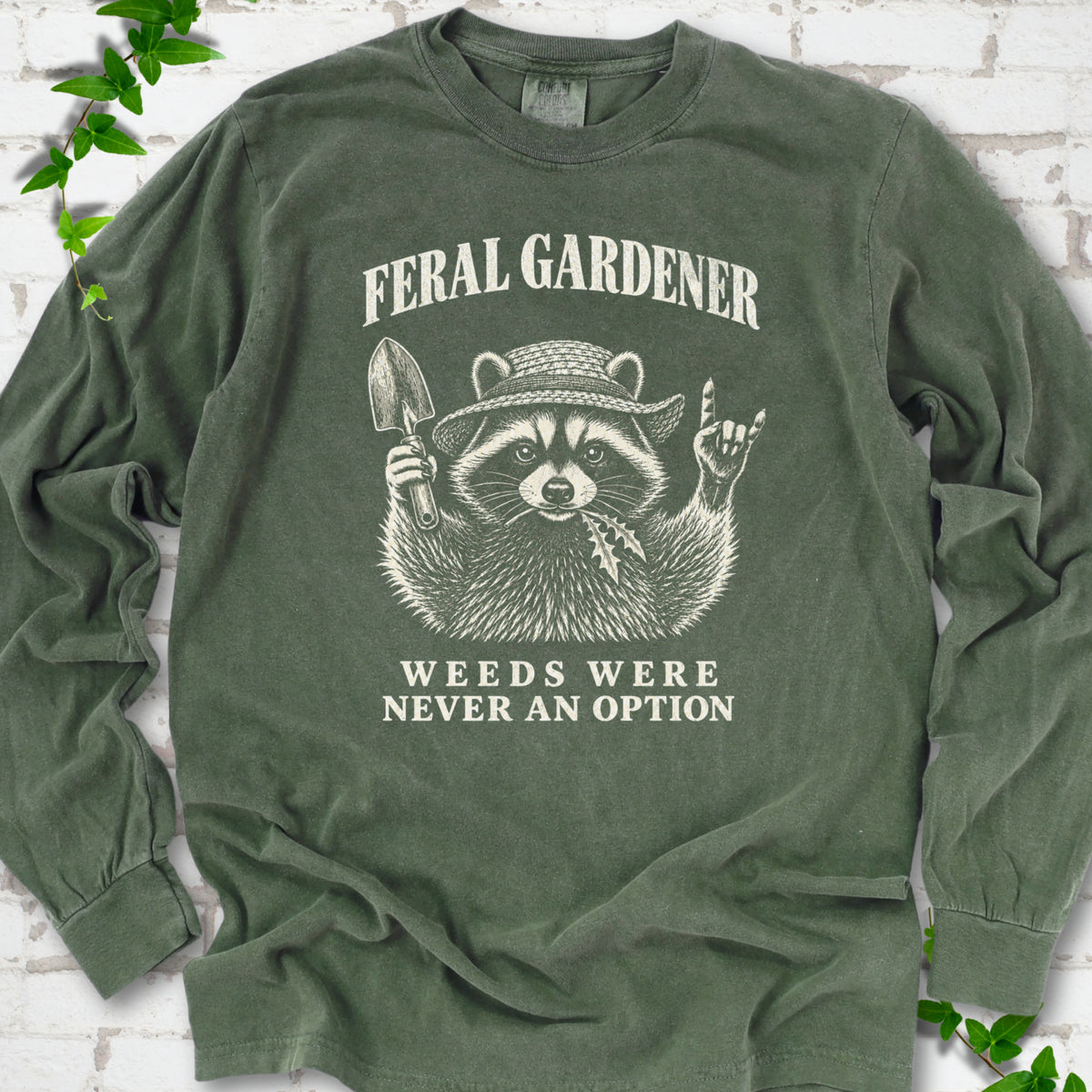 Hemp long sleeve tee laid flat, showing a cream raccoon in a straw hat holding a trowel, with "Feral Gardener" and "Weeds Were Never an Option" lettering.