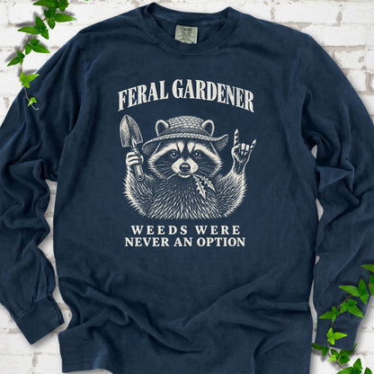 Midnight long sleeve tee laid flat, showing a cream raccoon in a straw hat holding a trowel, with "Feral Gardener" and "Weeds Were Never an Option" lettering.