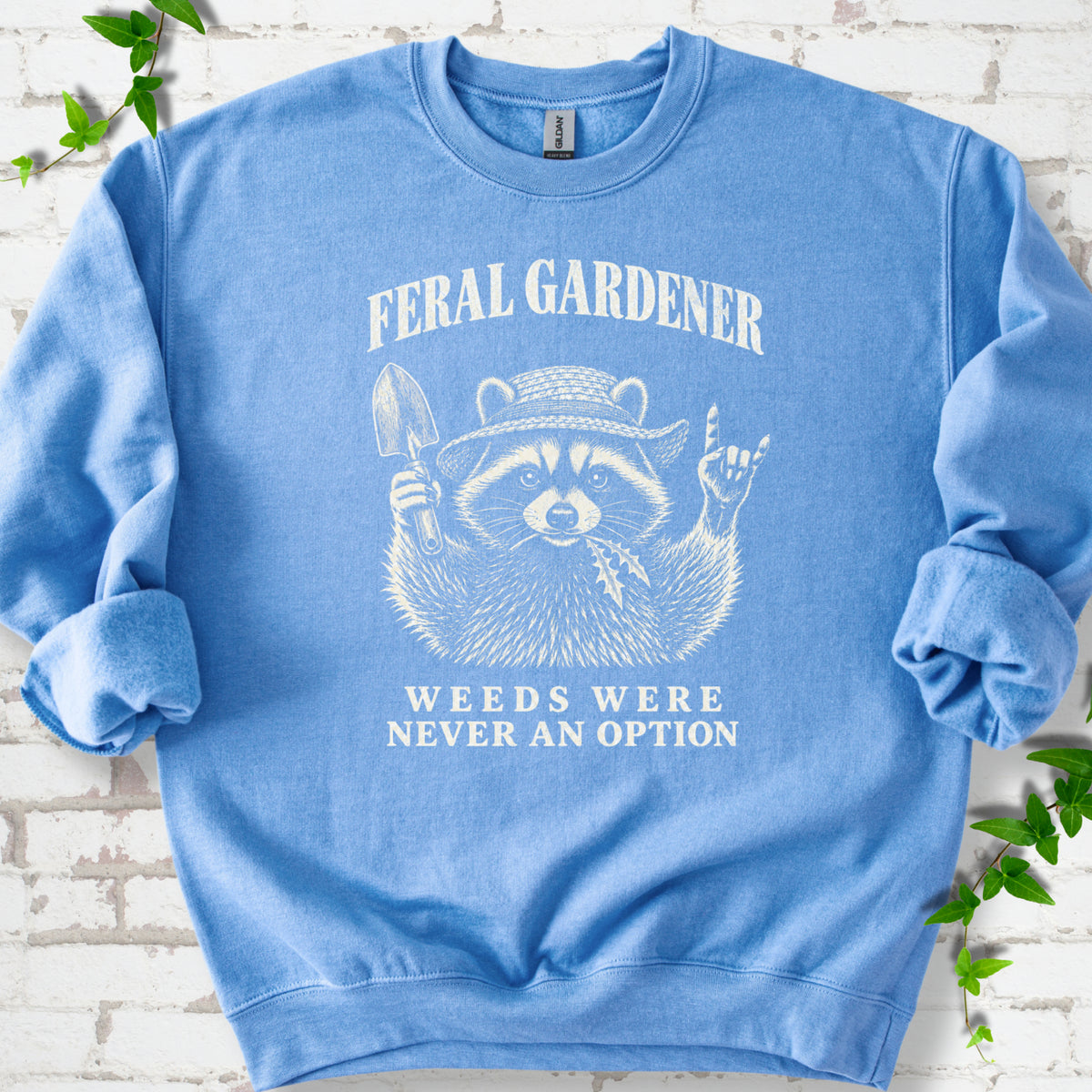 Carolina Blue classic sweatshirt laid flat, showing a cream raccoon in a straw hat holding a trowel, with "Feral Gardener" and "Weeds Were Never an Option" lettering.