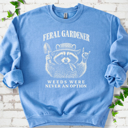Carolina Blue classic sweatshirt laid flat, showing a cream raccoon in a straw hat holding a trowel, with "Feral Gardener" and "Weeds Were Never an Option" lettering.