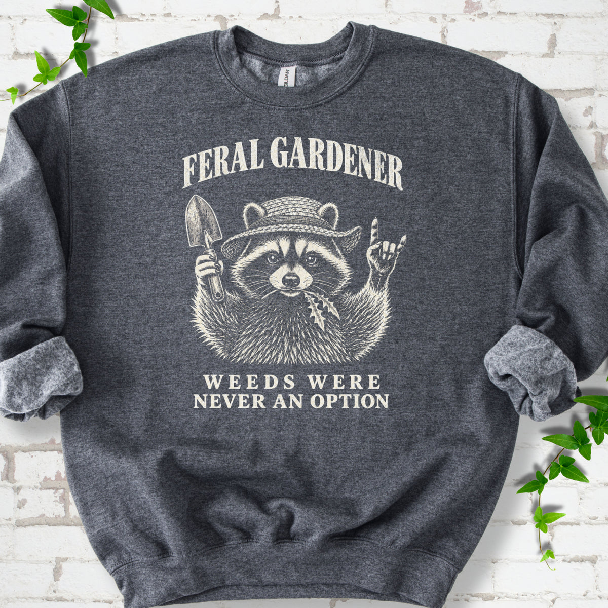 Dark Heather classic sweatshirt laid flat, showing a cream raccoon in a straw hat holding a trowel, with "Feral Gardener" and "Weeds Were Never an Option" lettering.