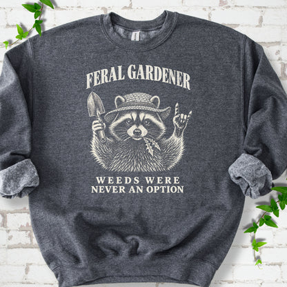 Dark Heather classic sweatshirt laid flat, showing a cream raccoon in a straw hat holding a trowel, with "Feral Gardener" and "Weeds Were Never an Option" lettering.