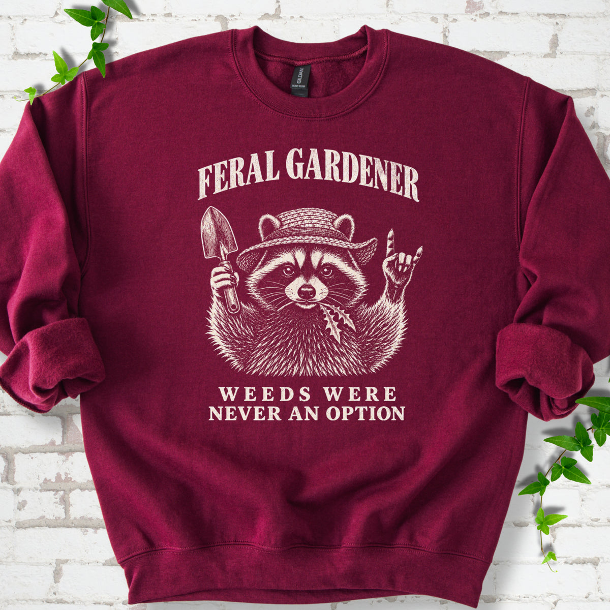 Maroon classic sweatshirt laid flat, showing a cream raccoon in a straw hat holding a trowel, with "Feral Gardener" and "Weeds Were Never an Option" lettering.