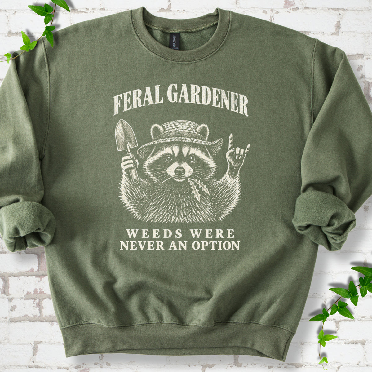 Military Green classic sweatshirt laid flat, showing a cream raccoon in a straw hat holding a trowel, with "Feral Gardener" and "Weeds Were Never an Option" lettering.